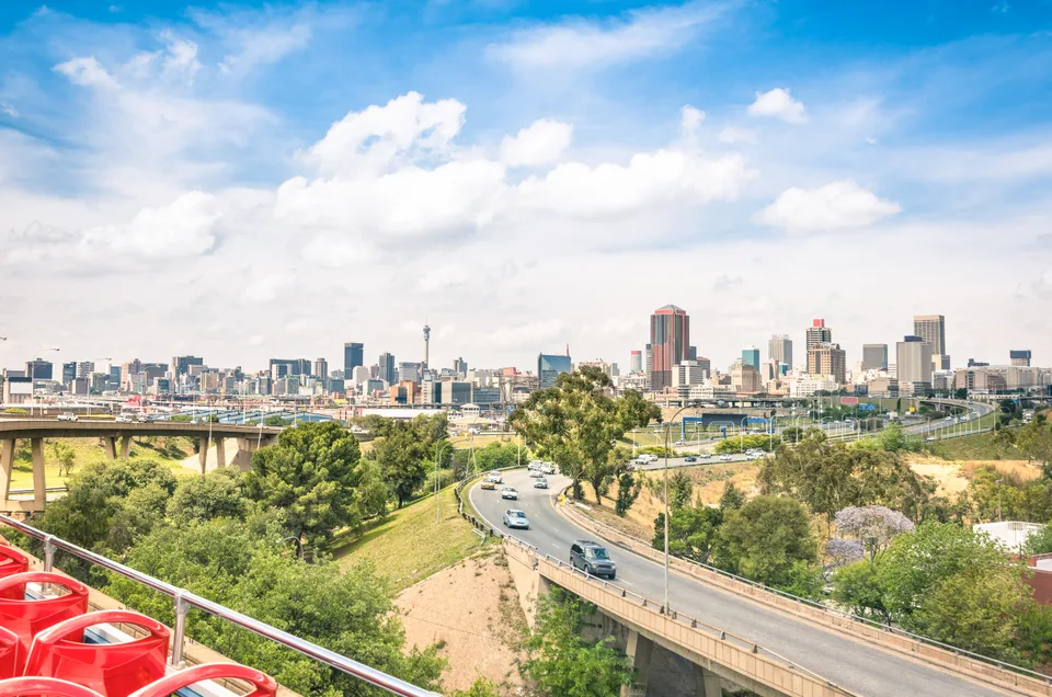 Johannesburg skyline beyond curving roads and green vegetation under a partly cloudy sky